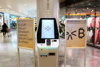 A cryptocurrency ATM is positioned in a modern shopping mall, flanked by two informational signs. The left sign lists currencies available for transaction including Bitcoin, Ethereum, Litecoin, and others, with a company logo at the bottom. The right sign invites users to exchange crypto, displaying symbols and a company logo. Shopper moving with a cart is visible in the background, along with clothing stores.