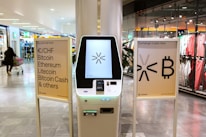 A cryptocurrency ATM is positioned in a modern shopping mall, flanked by two informational signs. The left sign lists currencies available for transaction including Bitcoin, Ethereum, Litecoin, and others, with a company logo at the bottom. The right sign invites users to exchange crypto, displaying symbols and a company logo. Shopper moving with a cart is visible in the background, along with clothing stores.