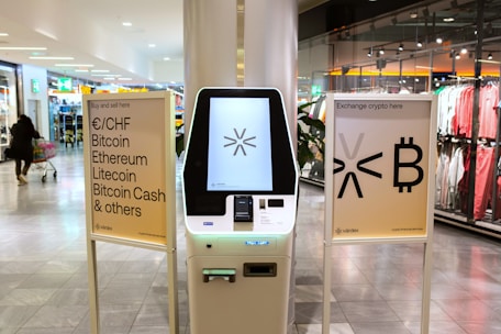 A cryptocurrency ATM is positioned in a modern shopping mall, flanked by two informational signs. The left sign lists currencies available for transaction including Bitcoin, Ethereum, Litecoin, and others, with a company logo at the bottom. The right sign invites users to exchange crypto, displaying symbols and a company logo. Shopper moving with a cart is visible in the background, along with clothing stores.