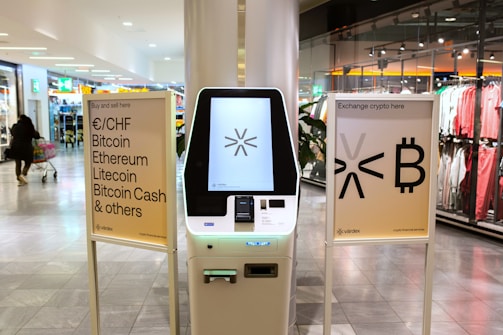 A cryptocurrency ATM is positioned in a modern shopping mall, flanked by two informational signs. The left sign lists currencies available for transaction including Bitcoin, Ethereum, Litecoin, and others, with a company logo at the bottom. The right sign invites users to exchange crypto, displaying symbols and a company logo. Shopper moving with a cart is visible in the background, along with clothing stores.