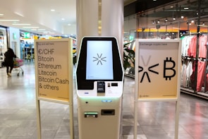 A cryptocurrency ATM is positioned in a modern shopping mall, flanked by two informational signs. The left sign lists currencies available for transaction including Bitcoin, Ethereum, Litecoin, and others, with a company logo at the bottom. The right sign invites users to exchange crypto, displaying symbols and a company logo. Shopper moving with a cart is visible in the background, along with clothing stores.