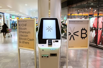 A cryptocurrency ATM is positioned in a modern shopping mall, flanked by two informational signs. The left sign lists currencies available for transaction including Bitcoin, Ethereum, Litecoin, and others, with a company logo at the bottom. The right sign invites users to exchange crypto, displaying symbols and a company logo. Shopper moving with a cart is visible in the background, along with clothing stores.