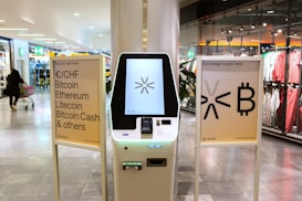 A cryptocurrency ATM is positioned in a modern shopping mall, flanked by two informational signs. The left sign lists currencies available for transaction including Bitcoin, Ethereum, Litecoin, and others, with a company logo at the bottom. The right sign invites users to exchange crypto, displaying symbols and a company logo. Shopper moving with a cart is visible in the background, along with clothing stores.