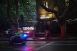 A dimly lit street scene with a highlighted sign reading 'BERNINI ART CAFE & RESTAURANT'. A motorcycle with a rider wearing a high-visibility jacket speeds past, while police lights flash in blue and red. Large trees and patio umbrellas surround the entrance of the brick building.