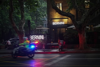 A dimly lit street scene with a highlighted sign reading 'BERNINI ART CAFE & RESTAURANT'. A motorcycle with a rider wearing a high-visibility jacket speeds past, while police lights flash in blue and red. Large trees and patio umbrellas surround the entrance of the brick building.