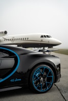 A luxury sports car with sleek black bodywork and blue accents is prominently featured in the foreground, showcasing its high-performance wheels. In the background, a private jet is parked on an overcast day, highlighting a scene of opulence and sophistication.