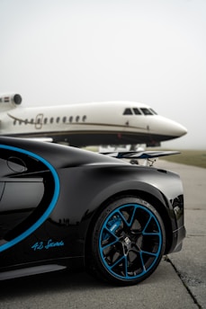 A luxury sports car with sleek black bodywork and blue accents is prominently featured in the foreground, showcasing its high-performance wheels. In the background, a private jet is parked on an overcast day, highlighting a scene of opulence and sophistication.