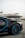 A luxury sports car with sleek black bodywork and blue accents is prominently featured in the foreground, showcasing its high-performance wheels. In the background, a private jet is parked on an overcast day, highlighting a scene of opulence and sophistication.