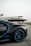 A luxury sports car with sleek black bodywork and blue accents is prominently featured in the foreground, showcasing its high-performance wheels. In the background, a private jet is parked on an overcast day, highlighting a scene of opulence and sophistication.