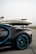 A luxury sports car with sleek black bodywork and blue accents is prominently featured in the foreground, showcasing its high-performance wheels. In the background, a private jet is parked on an overcast day, highlighting a scene of opulence and sophistication.