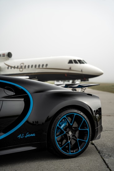 A luxury sports car with sleek black bodywork and blue accents is prominently featured in the foreground, showcasing its high-performance wheels. In the background, a private jet is parked on an overcast day, highlighting a scene of opulence and sophistication.