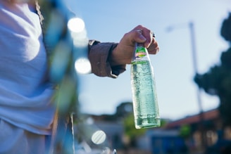 A hand holding a clear glass bottle with green apple soda against a blurred outdoor background. The person is wearing a checkered shirt, and the bright sunlight casts a natural reflection on the bottle.
