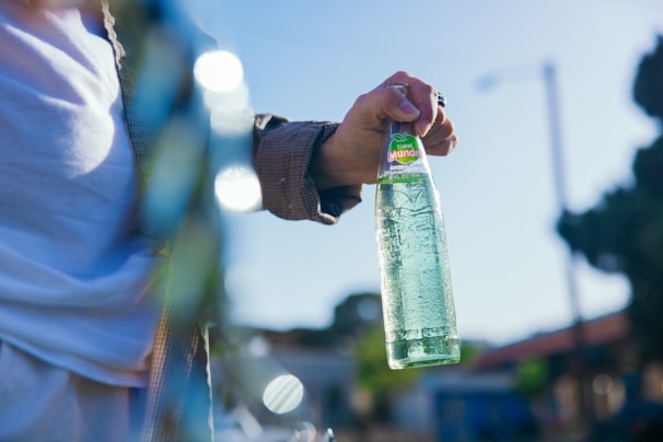A hand holding a clear glass bottle with green apple soda against a blurred outdoor background. The person is wearing a checkered shirt, and the bright sunlight casts a natural reflection on the bottle.