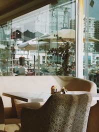 A cozy cafe setting with modern furniture, featuring a cushioned chair and a table with condiments. Large windows reveal an urban backdrop with buildings and a scattering of umbrellas, adding a touch of outdoor ambiance. The interior is warmly lit, offering a welcoming feel.