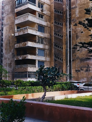 An old, weathered apartment building with multiple balconies and worn-down walls is seen. There are small plants and trees in front of the building, along with a neatly maintained green lawn. A portion of a parked car is visible in the background, next to the building. The overall appearance of the building suggests age and lack of recent maintenance.