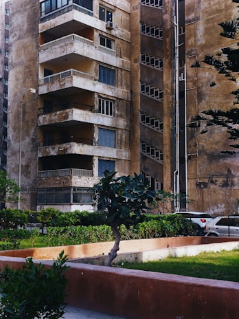 An old, weathered apartment building with multiple balconies and worn-down walls is seen. There are small plants and trees in front of the building, along with a neatly maintained green lawn. A portion of a parked car is visible in the background, next to the building. The overall appearance of the building suggests age and lack of recent maintenance.