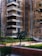 An old, weathered apartment building with multiple balconies and worn-down walls is seen. There are small plants and trees in front of the building, along with a neatly maintained green lawn. A portion of a parked car is visible in the background, next to the building. The overall appearance of the building suggests age and lack of recent maintenance.