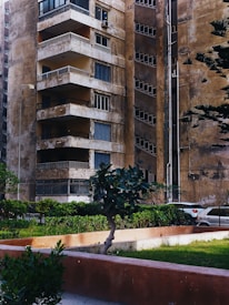 An old, weathered apartment building with multiple balconies and worn-down walls is seen. There are small plants and trees in front of the building, along with a neatly maintained green lawn. A portion of a parked car is visible in the background, next to the building. The overall appearance of the building suggests age and lack of recent maintenance.