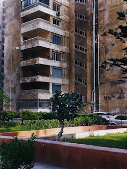 An old, weathered apartment building with multiple balconies and worn-down walls is seen. There are small plants and trees in front of the building, along with a neatly maintained green lawn. A portion of a parked car is visible in the background, next to the building. The overall appearance of the building suggests age and lack of recent maintenance.