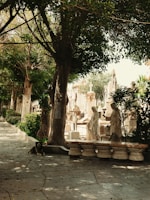 A peaceful cemetery scene with statues and tombstones bordered by a row of lush trees. Sunlight filters through the leaves, casting dappled shadows on the path. A dog lies calmly on the ground near a tree.