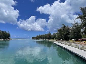 A serene waterfront landscape with calm turquoise waters reflecting a partly cloudy sky. Dense, lush green trees line the banks in the background, creating a natural border. A wooden dock runs along the right side, with a few parked vehicles visible behind it.