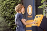 A woman using a Bitcoin ATM located outdoors. She is operating the touchscreen of the ATM while holding a smartphone in her other hand. The ATM and the surrounding area have a Bitcoin logo. The background includes a leafy green wall.