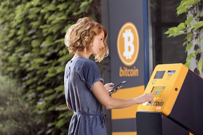 A woman using a Bitcoin ATM located outdoors. She is operating the touchscreen of the ATM while holding a smartphone in her other hand. The ATM and the surrounding area have a Bitcoin logo. The background includes a leafy green wall.
