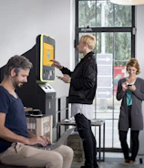 A modern interior with several people interacting with technology, including a digital currency ATM. One person is using a laptop at a table, another is standing at an ATM, and others are using their phones.