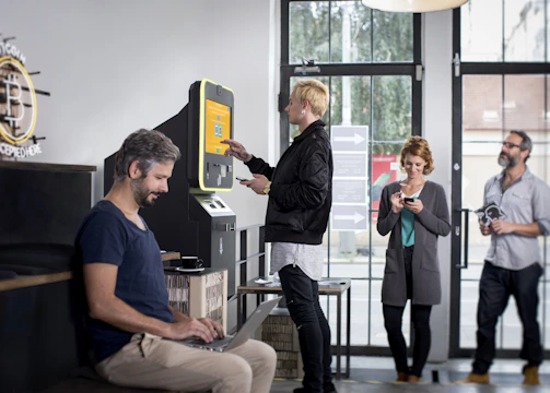 A modern interior with several people interacting with technology, including a digital currency ATM. One person is using a laptop at a table, another is standing at an ATM, and others are using their phones.