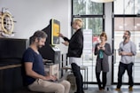 A modern interior with several people interacting with technology, including a digital currency ATM. One person is using a laptop at a table, another is standing at an ATM, and others are using their phones.