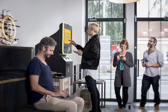 A modern interior with several people interacting with technology, including a digital currency ATM. One person is using a laptop at a table, another is standing at an ATM, and others are using their phones.