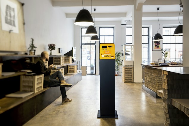 A modern interior space featuring a sleek, well-lit environment with high ceilings and pendant lights. In the center, there is a bright yellow Bitcoin ATM standing prominently on a black pedestal. To the left, a person is sitting on a bench with a cup, seemingly relaxed. On the right, a counter area displays plants and decorative items, with shelves made from wooden crates.