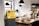 A stylish cafe interior features a yellow Bitcoin ATM positioned prominently. The counter is wooden and holds several glass domes with pastries inside. Modern pendant lights hang from the ceiling, and plants add a touch of green to the space.