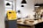 A stylish cafe interior features a yellow Bitcoin ATM positioned prominently. The counter is wooden and holds several glass domes with pastries inside. Modern pendant lights hang from the ceiling, and plants add a touch of green to the space.