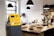 A stylish cafe interior features a yellow Bitcoin ATM positioned prominently. The counter is wooden and holds several glass domes with pastries inside. Modern pendant lights hang from the ceiling, and plants add a touch of green to the space.