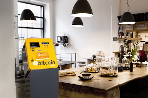 A stylish cafe interior features a yellow Bitcoin ATM positioned prominently. The counter is wooden and holds several glass domes with pastries inside. Modern pendant lights hang from the ceiling, and plants add a touch of green to the space.