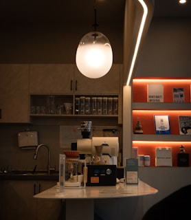 A cozy coffee shop interior featuring a modern light fixture hanging above a coffee-making station with a coffee grinder and brewing equipment. The counter is adorned with various containers and a small sign. On the wall, shelves display books, decorations, and a miniature Christmas tree, with soft lighting creating a warm atmosphere. A sink and kitchen cabinets are visible in the background.