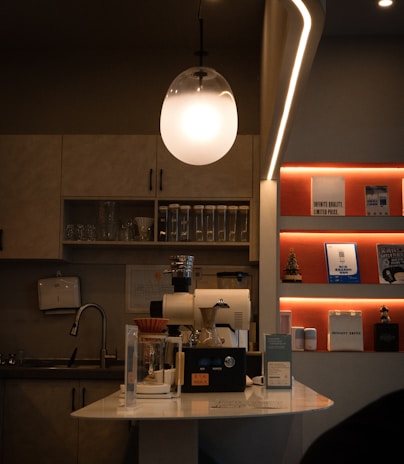 A cozy coffee shop interior featuring a modern light fixture hanging above a coffee-making station with a coffee grinder and brewing equipment. The counter is adorned with various containers and a small sign. On the wall, shelves display books, decorations, and a miniature Christmas tree, with soft lighting creating a warm atmosphere. A sink and kitchen cabinets are visible in the background.