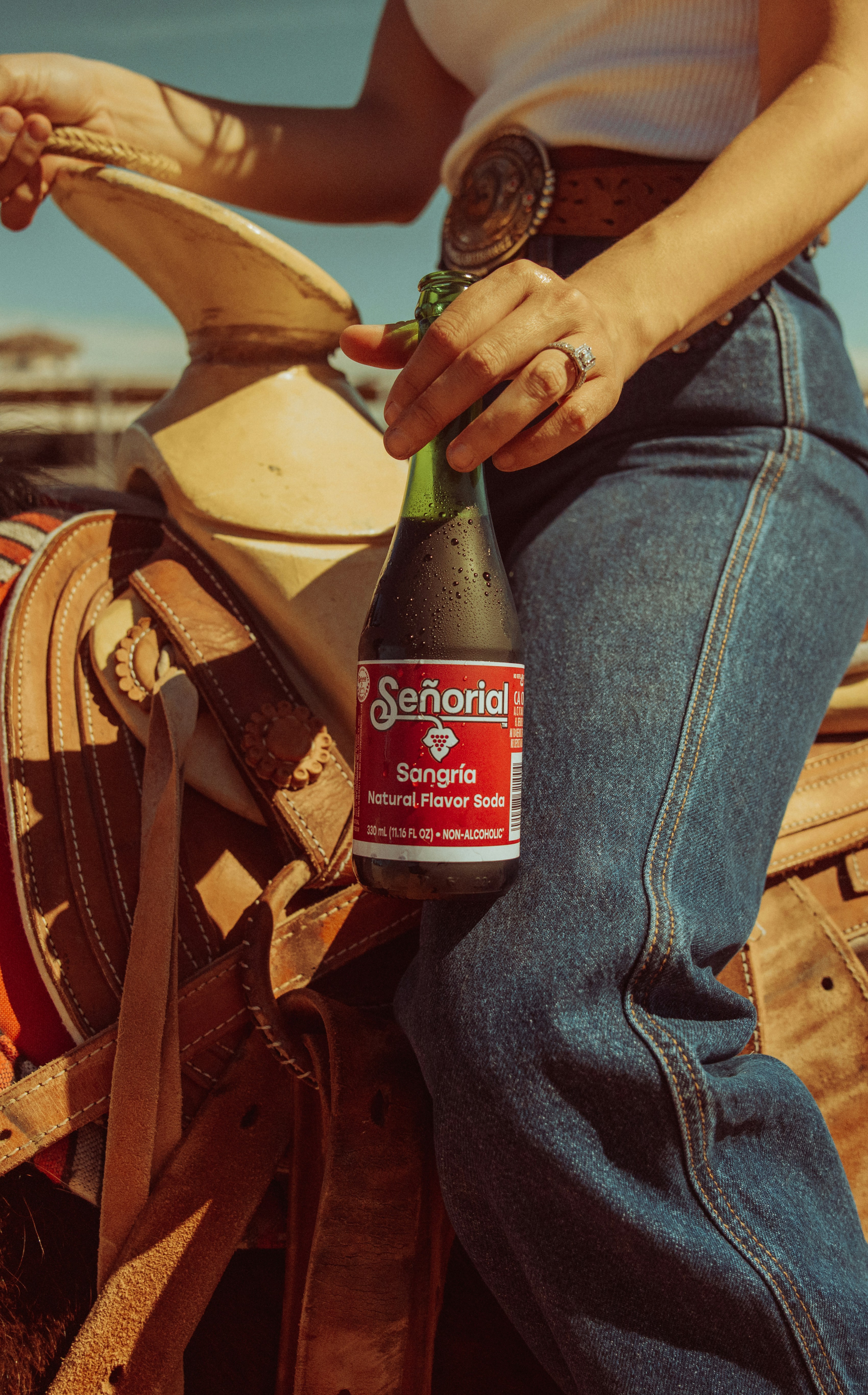 Photograph of a Señorial Sangria soda bottle held near a leather saddlebag and denim jeans in bright outdoor light.