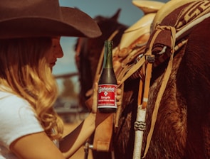 A person, wearing a cowboy hat and a white shirt, is holding a bottle of Senorial Sangria flavored soda while standing next to a saddled horse. The scene has a rustic, Western vibe with warm sunlight casting a golden hue over the setting.