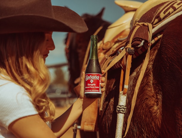 A person, wearing a cowboy hat and a white shirt, is holding a bottle of Senorial Sangria flavored soda while standing next to a saddled horse. The scene has a rustic, Western vibe with warm sunlight casting a golden hue over the setting.