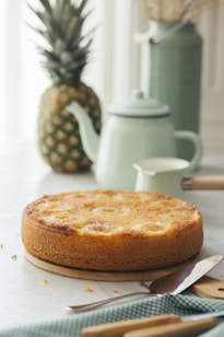 A freshly baked pineapple upside-down cake is placed on a circular wooden board, enticing with its golden-brown crust and visible fruit pieces. In the background, a large ripe pineapple stands alongside a mint green teapot and a matching milk jug, creating a cozy and inviting scene. The table is adorned with a checkered cloth, and a knife rests beside the cake, hinting at a soon-to-be-served dessert.