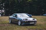 A classic BMW car is parked on a grassy field, with trees in the background. The car appears to be well-maintained and polished, reflecting light on its blue surface. There are a few more vehicles visible in the background.