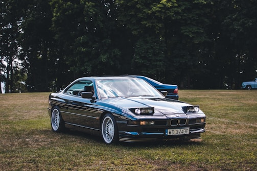 A classic BMW car is parked on a grassy field, with trees in the background. The car appears to be well-maintained and polished, reflecting light on its blue surface. There are a few more vehicles visible in the background.