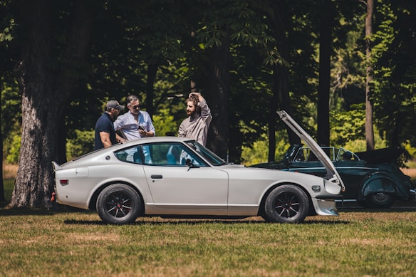 A classic white sports car is parked on grass with its hood open, surrounded by three men who appear to be inspecting or discussing it. The scene takes place in a wooded area with tall trees providing shade. Another vintage car, partially visible, is parked nearby.
