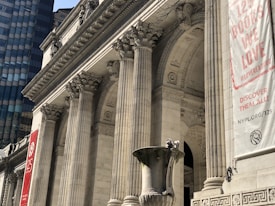A grand architectural structure featuring large Corinthian columns and intricate stone carvings. The building's facade showcases classical design elements with decorative reliefs. A large banner hangs on the right side, displaying text about books and a logo. In the foreground, there is an ornate stone urn.