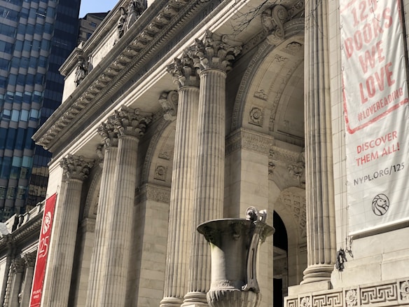 A grand architectural structure featuring large Corinthian columns and intricate stone carvings. The building's facade showcases classical design elements with decorative reliefs. A large banner hangs on the right side, displaying text about books and a logo. In the foreground, there is an ornate stone urn.