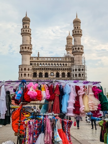 A bustling market stall is set up in front of a historic monument with four ornate minarets. The stall displays a vibrant array of colorful, frilly dresses hanging from a canopy-covered rack. People are seen walking around, adding to the lively atmosphere.