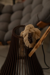 A thick, braided rope is tied around the top of a dark, metal cage-like structure. The background features a blurred, cushion-like texture in shades of grey and brown.
