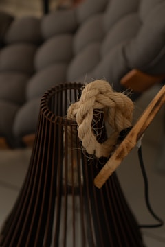 A thick, braided rope is tied around the top of a dark, metal cage-like structure. The background features a blurred, cushion-like texture in shades of grey and brown.